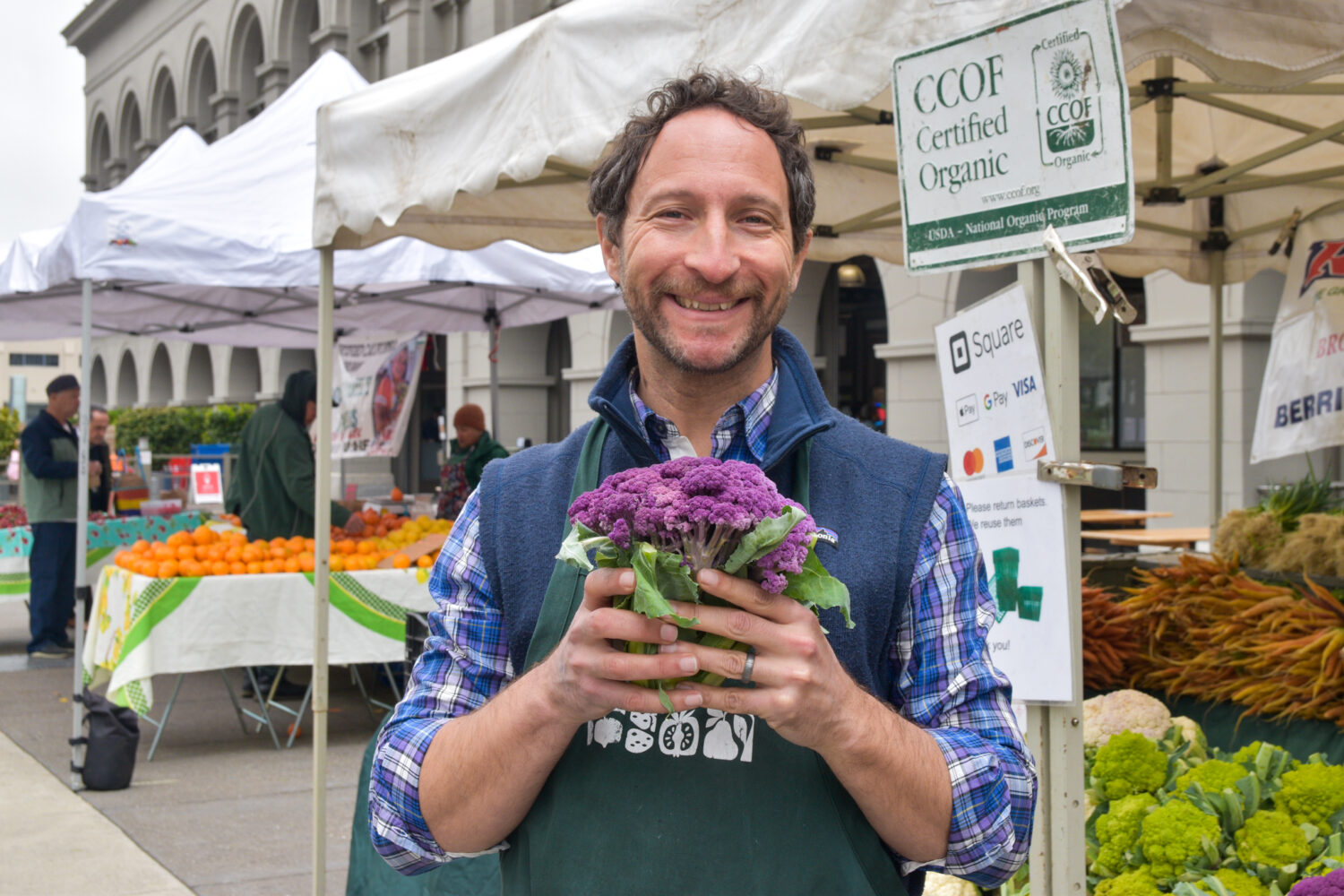 Richard Bernstein holds a purple cauliflower in front of farmers market tents