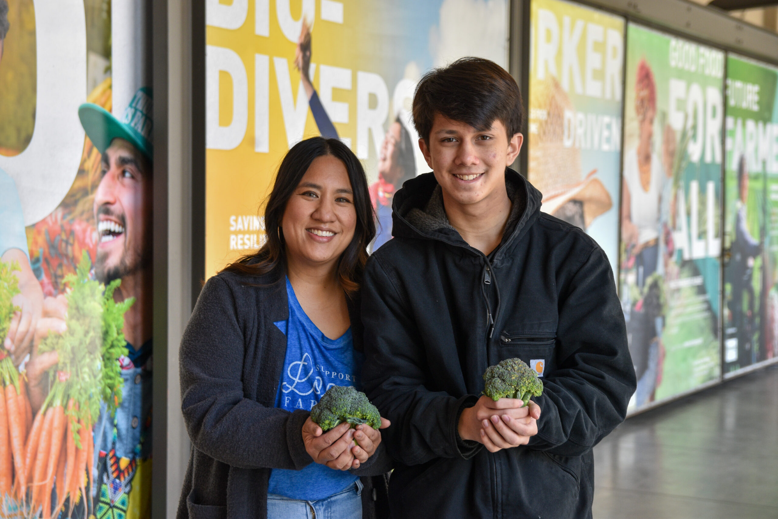Leslie and Josiah, holding vegetables, in front of The Food Change photo murals